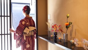 Une personne vêtue d’un costume traditionnel, debout près d’une fenêtre donnant sur un paysage.