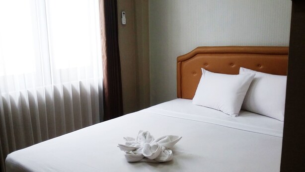 A neatly made bed with white linens and pillows, a wooden headboard, and a folded white towel on the bed. - DeKopen Rooms & Convention (Malang)