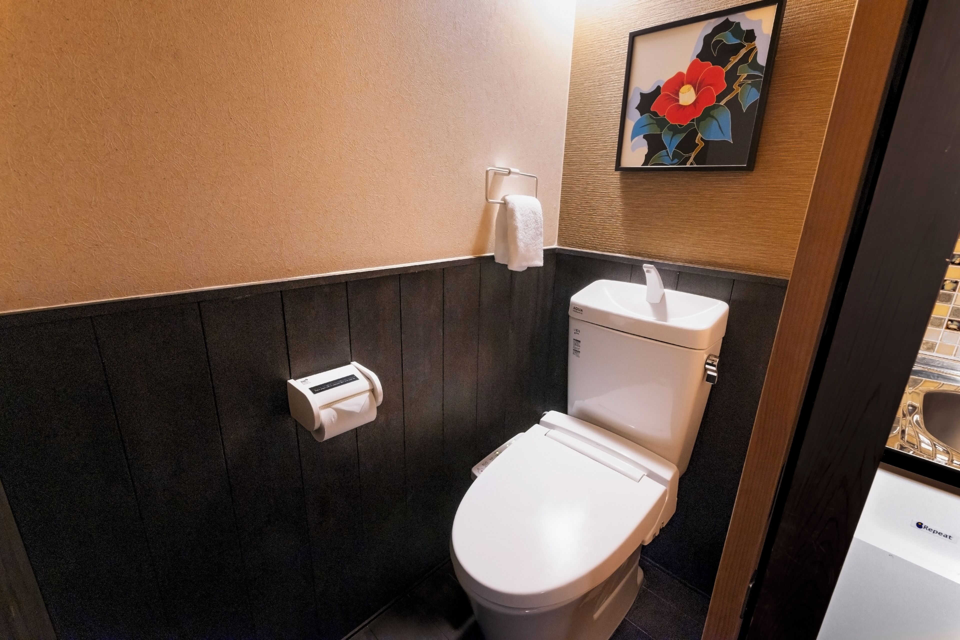 A compact bathroom with a white toilet, a paper towel dispenser, and a framed floral artwork on the wall.