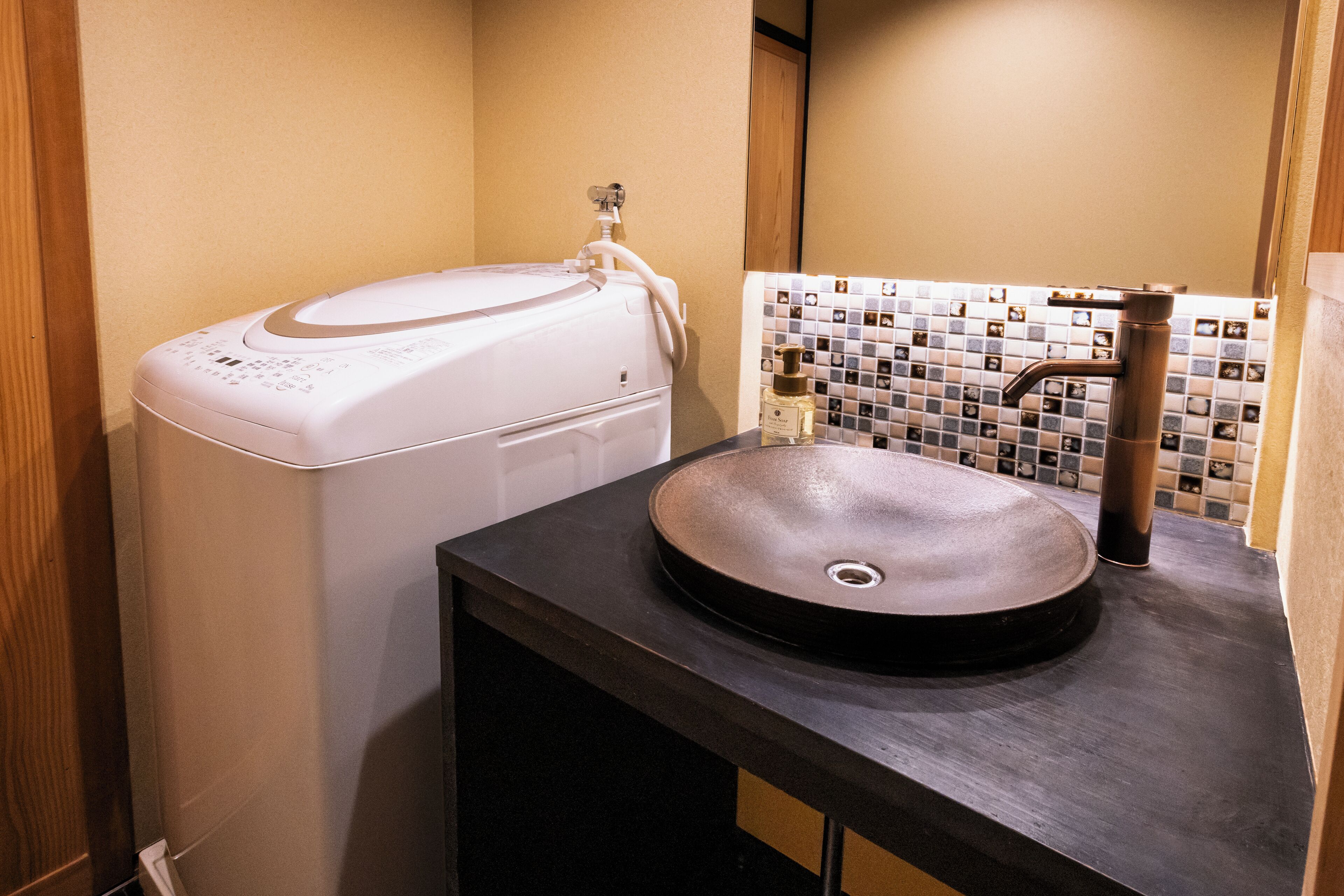 Un baño moderno con lavadora integrada, un lavamanos redondo con grifo de cobre y un respaldo de azulejos.
