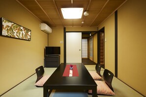 Traditional Japanese-style room with tatami flooring, featuring a central table and chairs, mounted TV, and sliding door to a separate room.