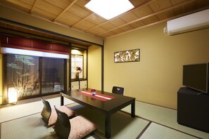 Experience the tranquility of a traditional Japanese-style room with tatami flooring and a low table, featuring a mounted television and a sliding door leading to a serene garden view.