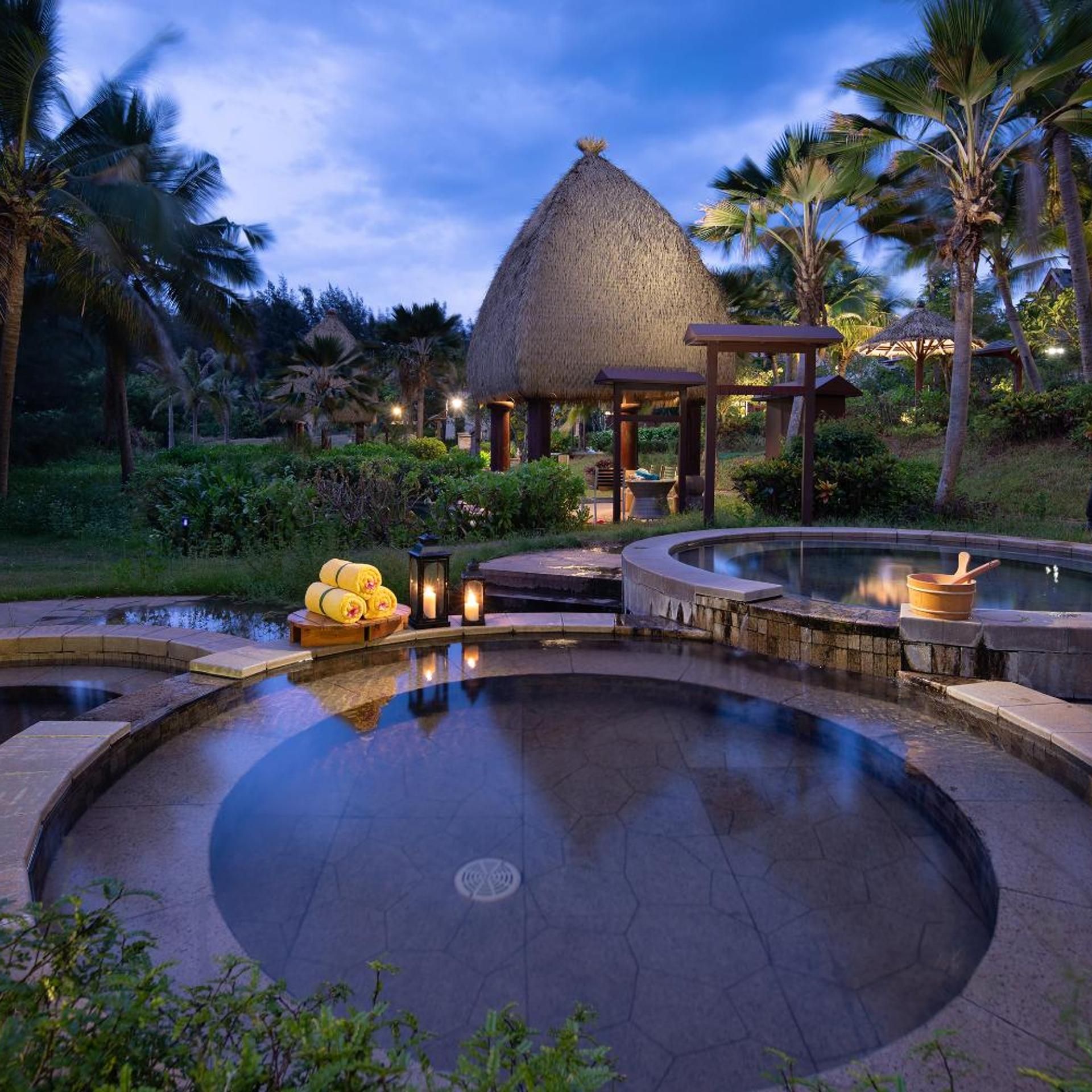 A resort pool area with a thatched-roof structure, surrounded by palm trees and greenery.