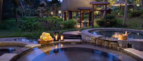 A resort pool area with a thatched-roof structure, surrounded by palm trees and greenery.