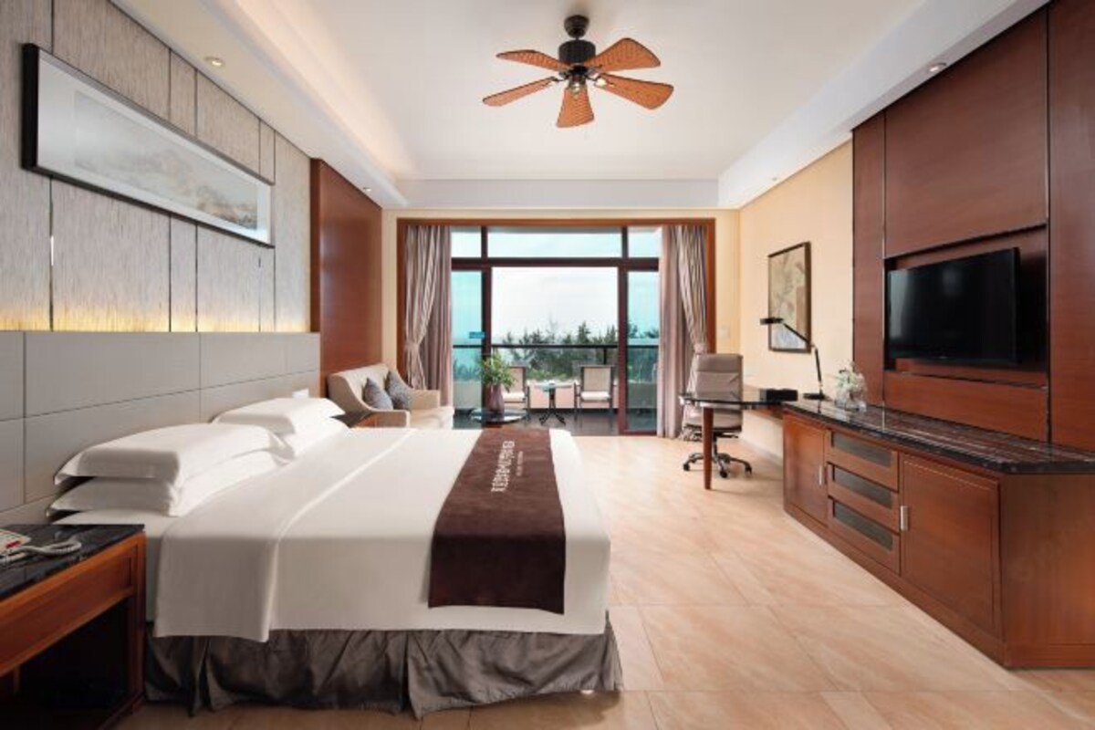 A hotel room with a large bed, a desk with a TV, a balcony with a view, and a ceiling fan.