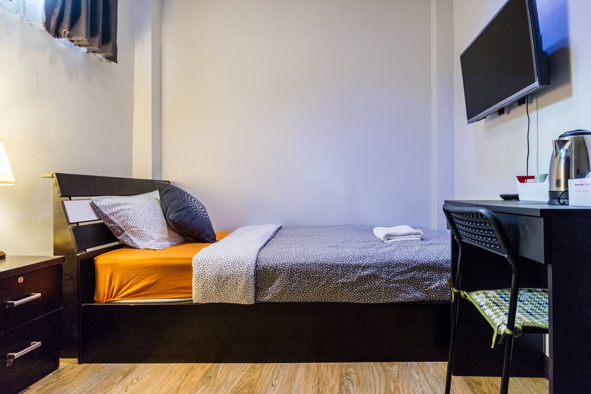 A single bed with an orange mattress, a black headboard, and two pillows. A desk with a chair is to the right, and a flat-screen TV is mounted on the wall above it.
