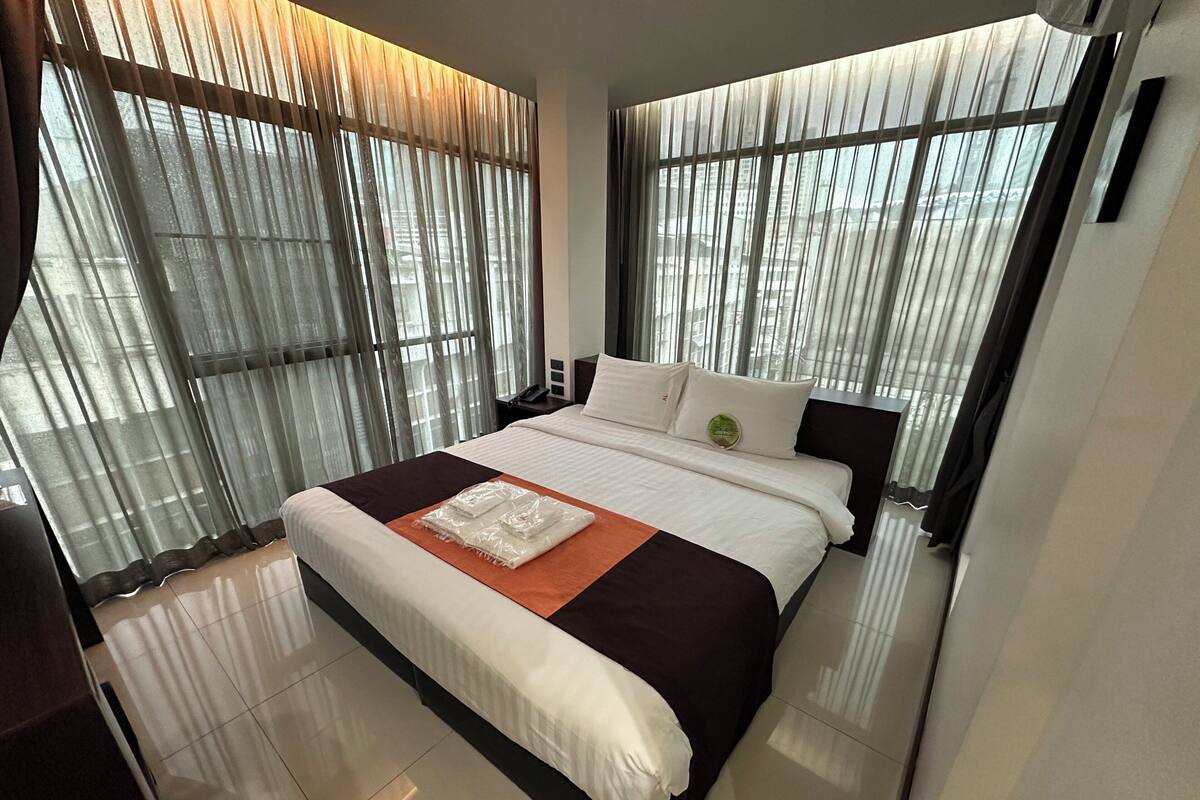 Ein modernes Hotelzimmer mit einem großen Bett, einem Nachttisch und Blick durch leichte Vorhänge.