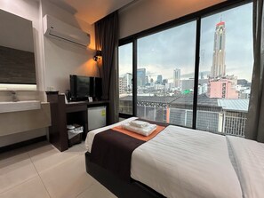 A hotel room with a bed, a desk with a TV, a mini-fridge, a bathroom with a sink, and a large window with a city view. - Collection O The Bangkok Airport Link Suite (Bangkok)