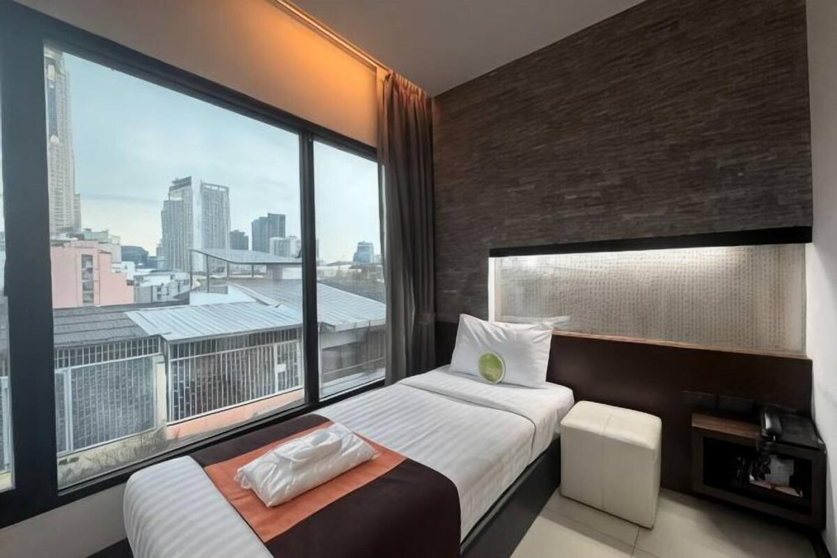 Ein Hotelzimmer mit großem Fenster, einem Bett, einem Nachttisch und Blick auf die Stadt.