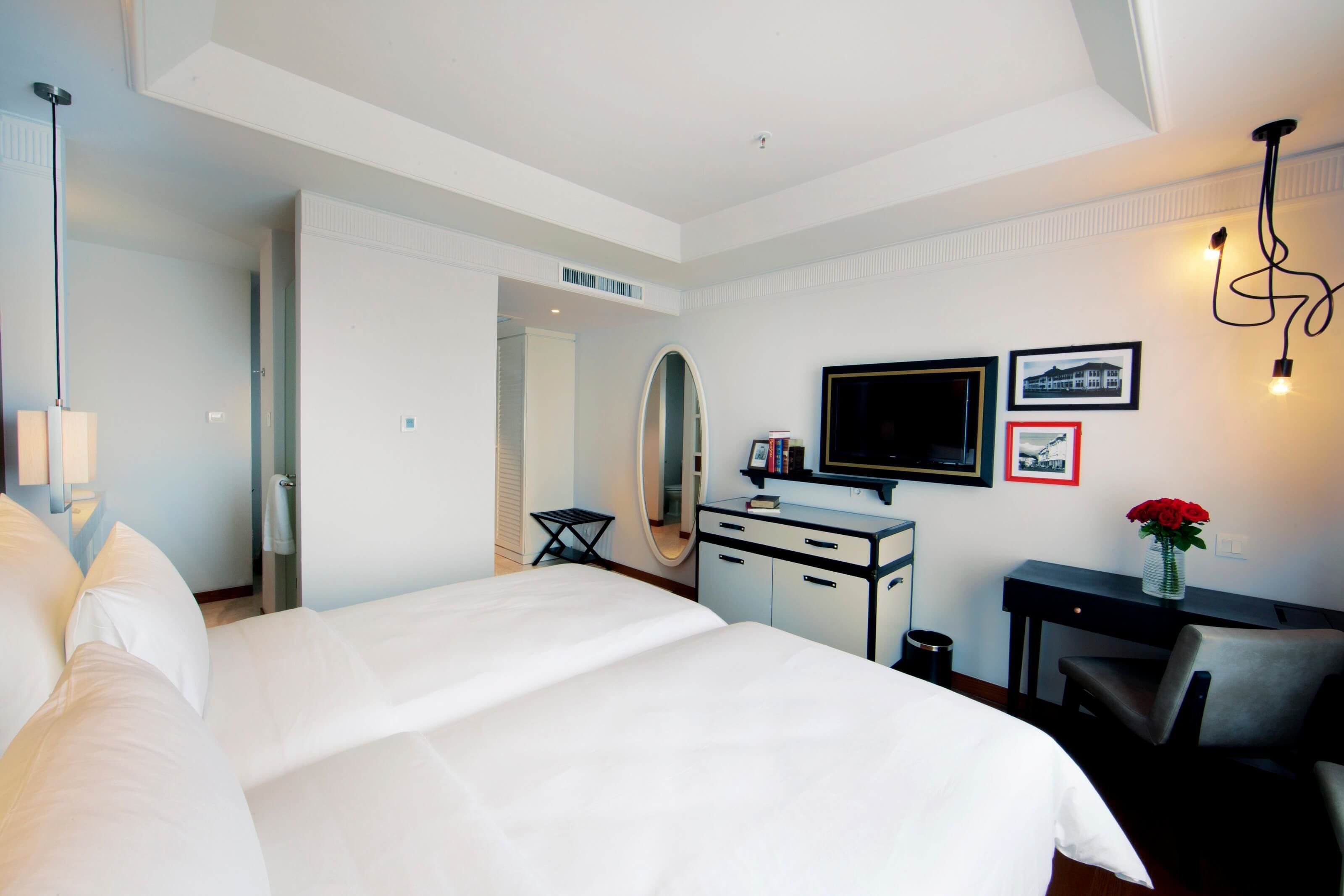 A hotel room with a large bed, a desk with a chair, a television, a mirror, and a small table with a vase of flowers.