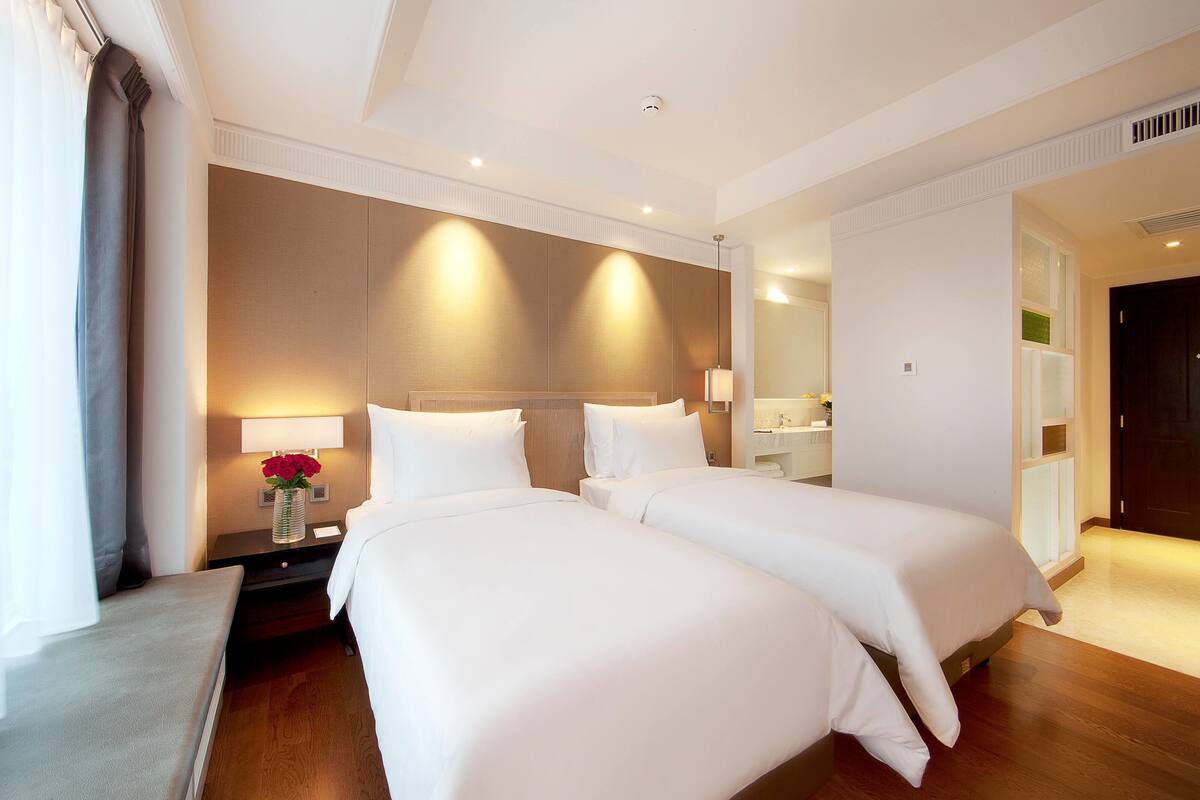 Habitación de hotel con dos camas, una mesita de noche con un jarrón de flores, un espejo y un baño visible a través de una puerta abierta.