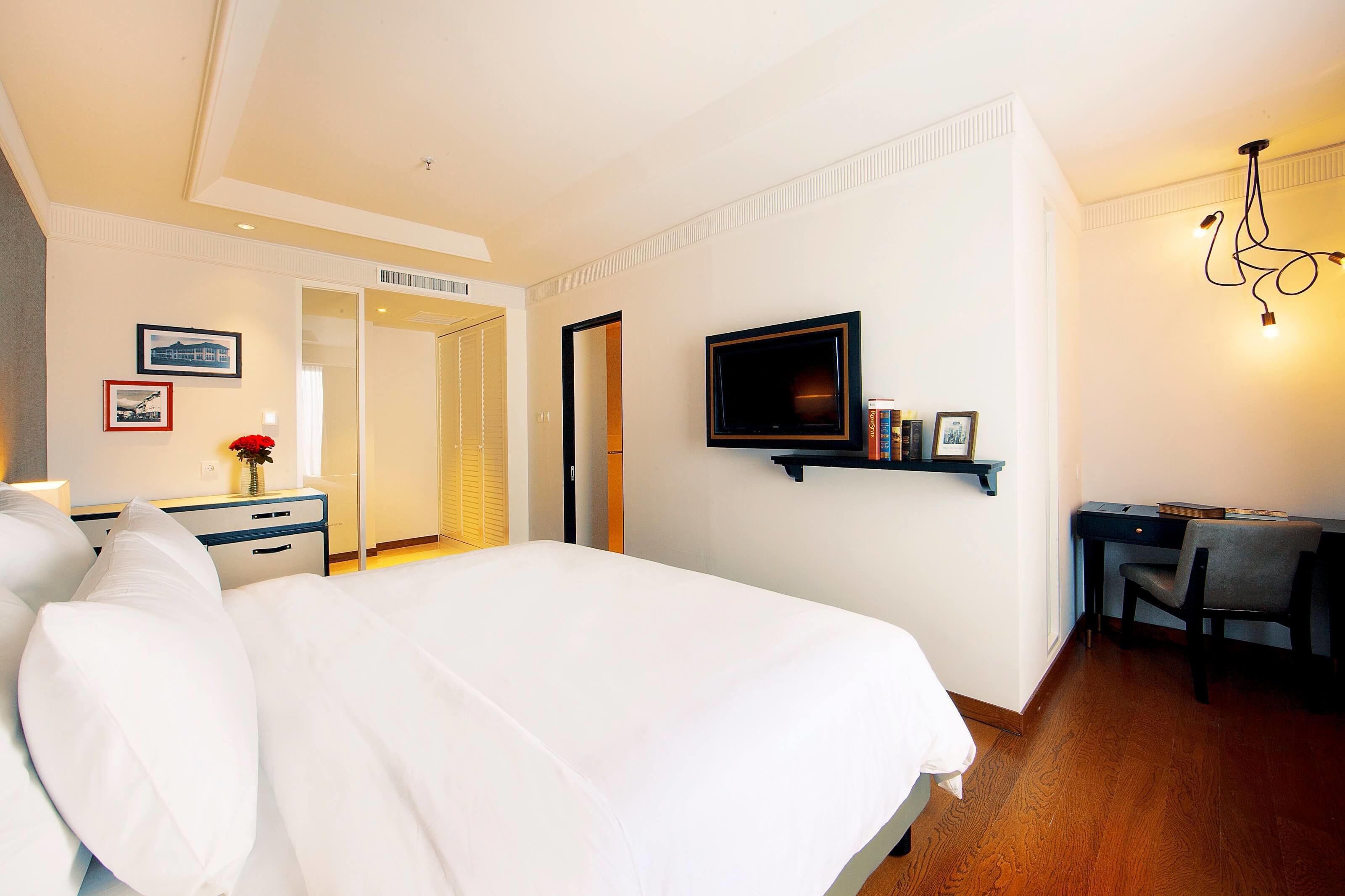 A hotel room with a large bed, a desk with a chair, a TV mounted on the wall, and a door leading to another room.