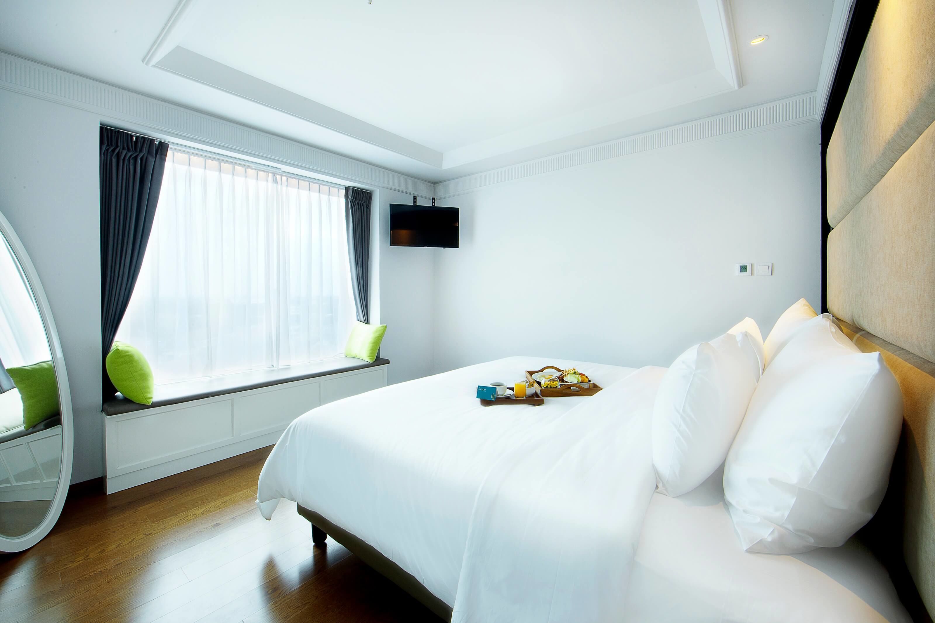 Ein Hotelzimmer mit einem großen Bett, einem Fenster mit Vorhängen, einem an der Wand montierten Flachbildfernseher und einem Nachttisch mit einer Lampe.