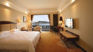 A hotel room with a large bed, a desk, a chair, and a TV. There is a balcony with a view of the water and hills. - Anqing Country Garden Phoenix Hotel (Anqing)