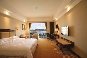 Quarto de hotel com uma cama grande, uma mesa, uma cadeira e uma TV. Há uma varanda com vista para a água e colinas.