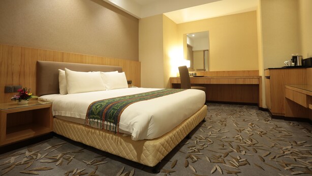 A hotel room with a large bed, a wooden headboard, a bedside table with a lamp, a mirror, and a bathroom area with a sink and toilet. - Imperial Riverbank Hotel Kuching (Kuching)