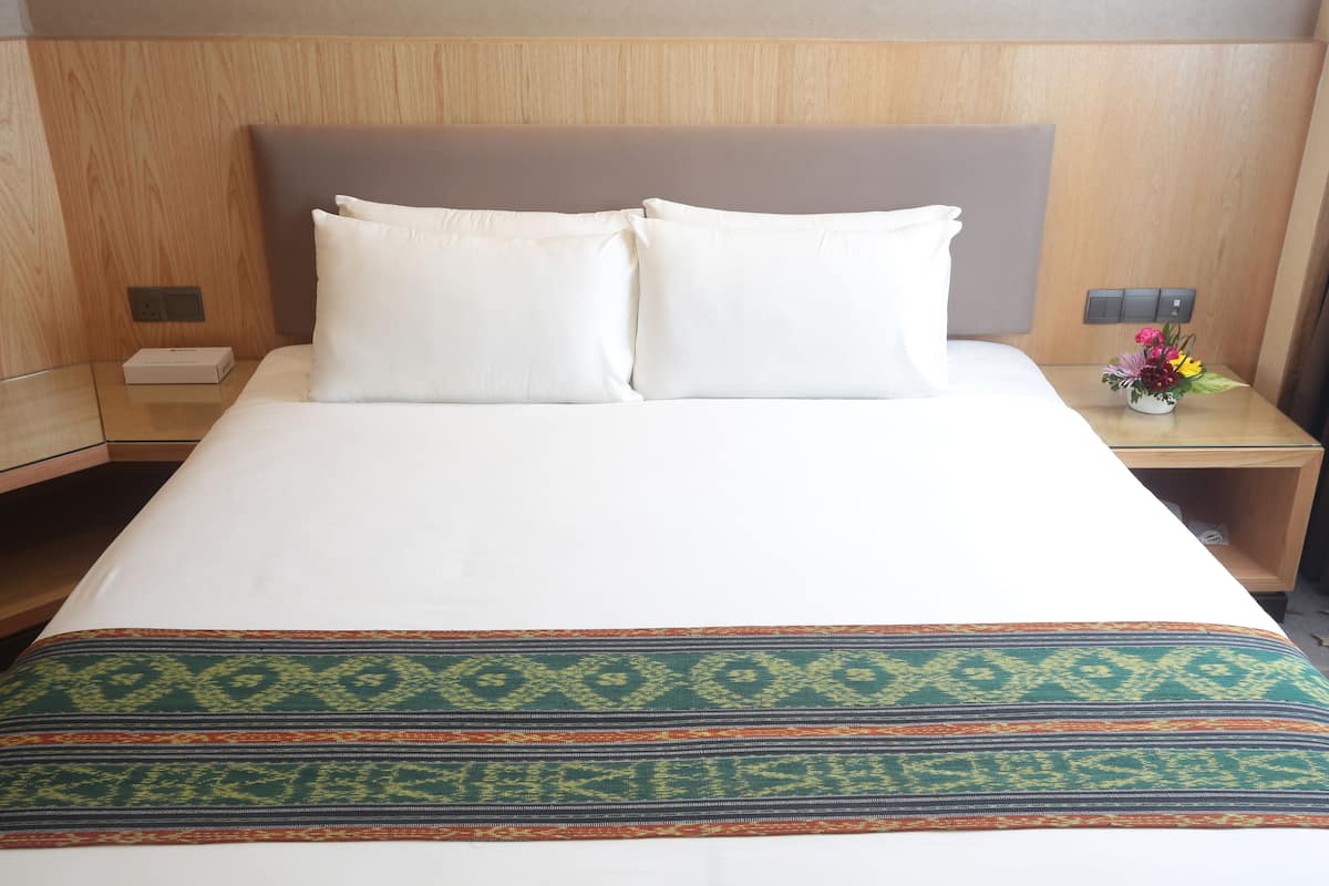Ein Hotelzimmer mit einem Bett, zwei Kissen, einem Nachttisch mit Blumenvase und einem Holz-Kopfteil.