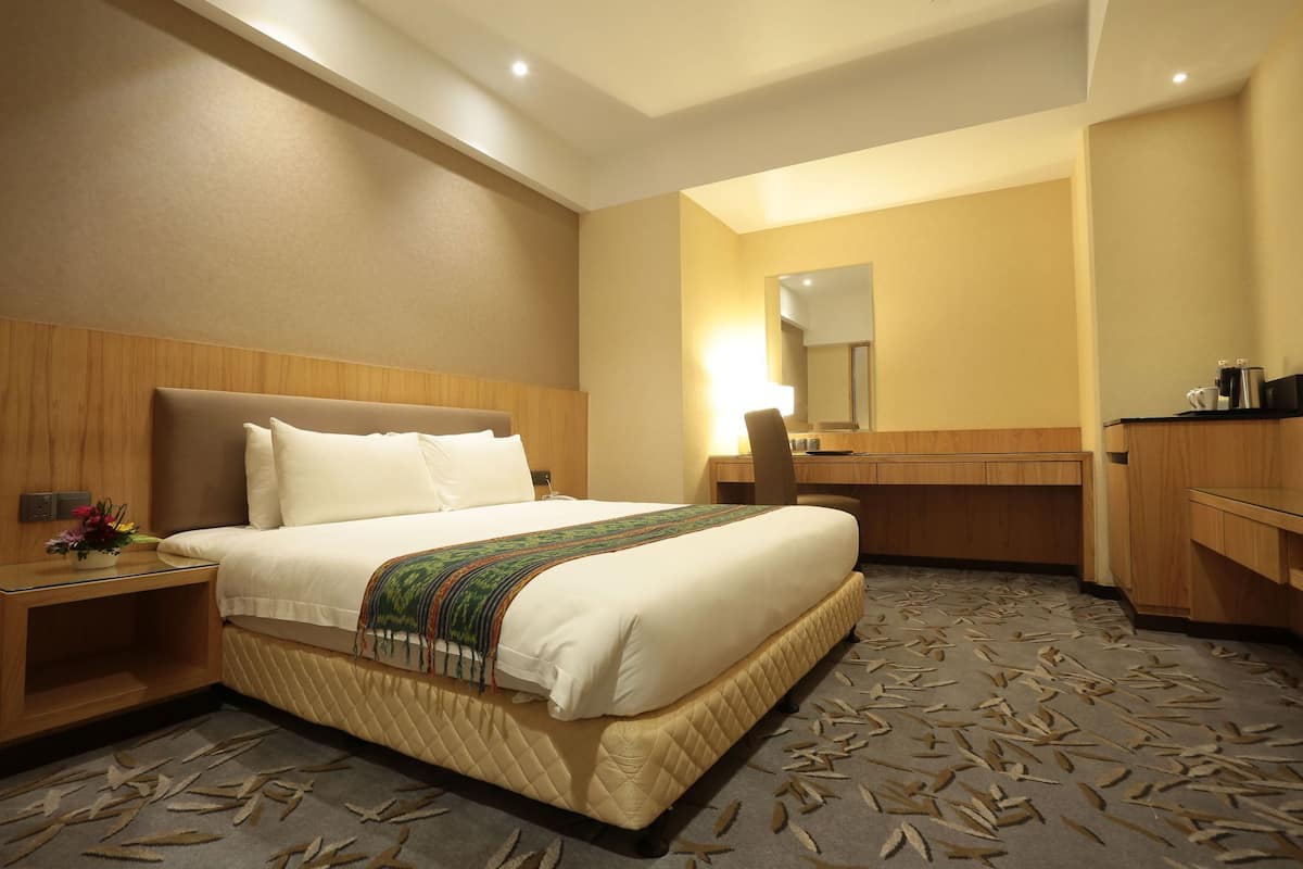 Ein Hotelzimmer mit einem großen Bett, einem Nachttisch mit Lampe, einem Spiegel, einem Schreibtisch und einer Küchenzeile.
