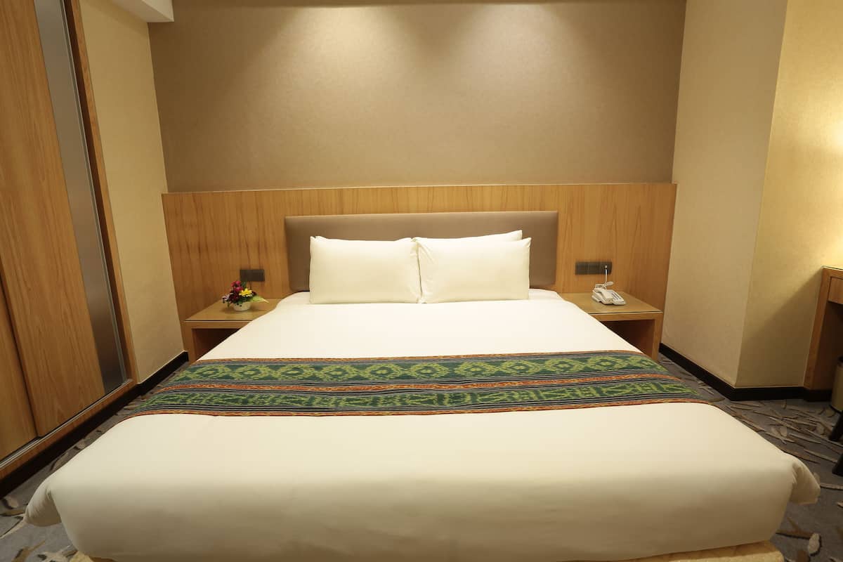 Ein Hotelzimmer mit einem großen Bett, einem Holz-Kopfteil, zwei Nachttischen mit Lampen sowie einer Blumenkomposition auf einem Tisch.