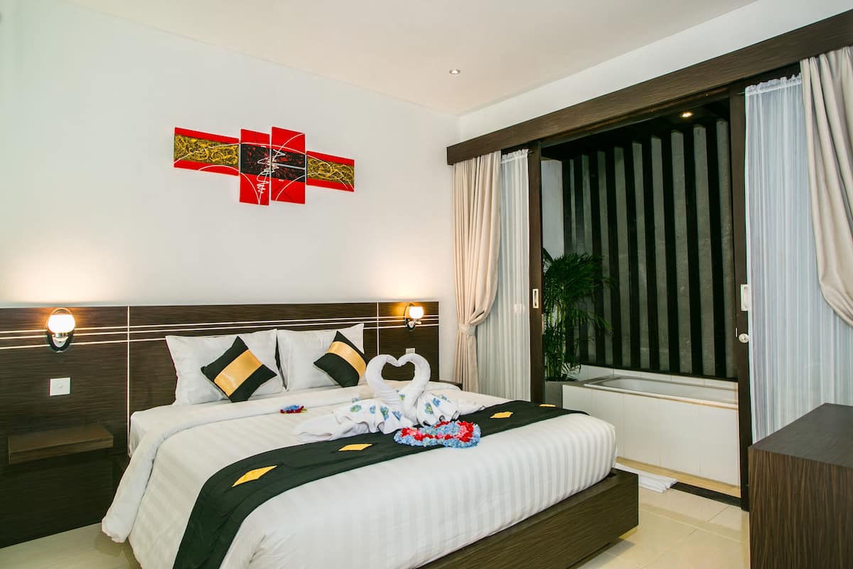 A hotel room with a large bed, a headboard with light fixtures, a window with curtains, and a wall art piece.