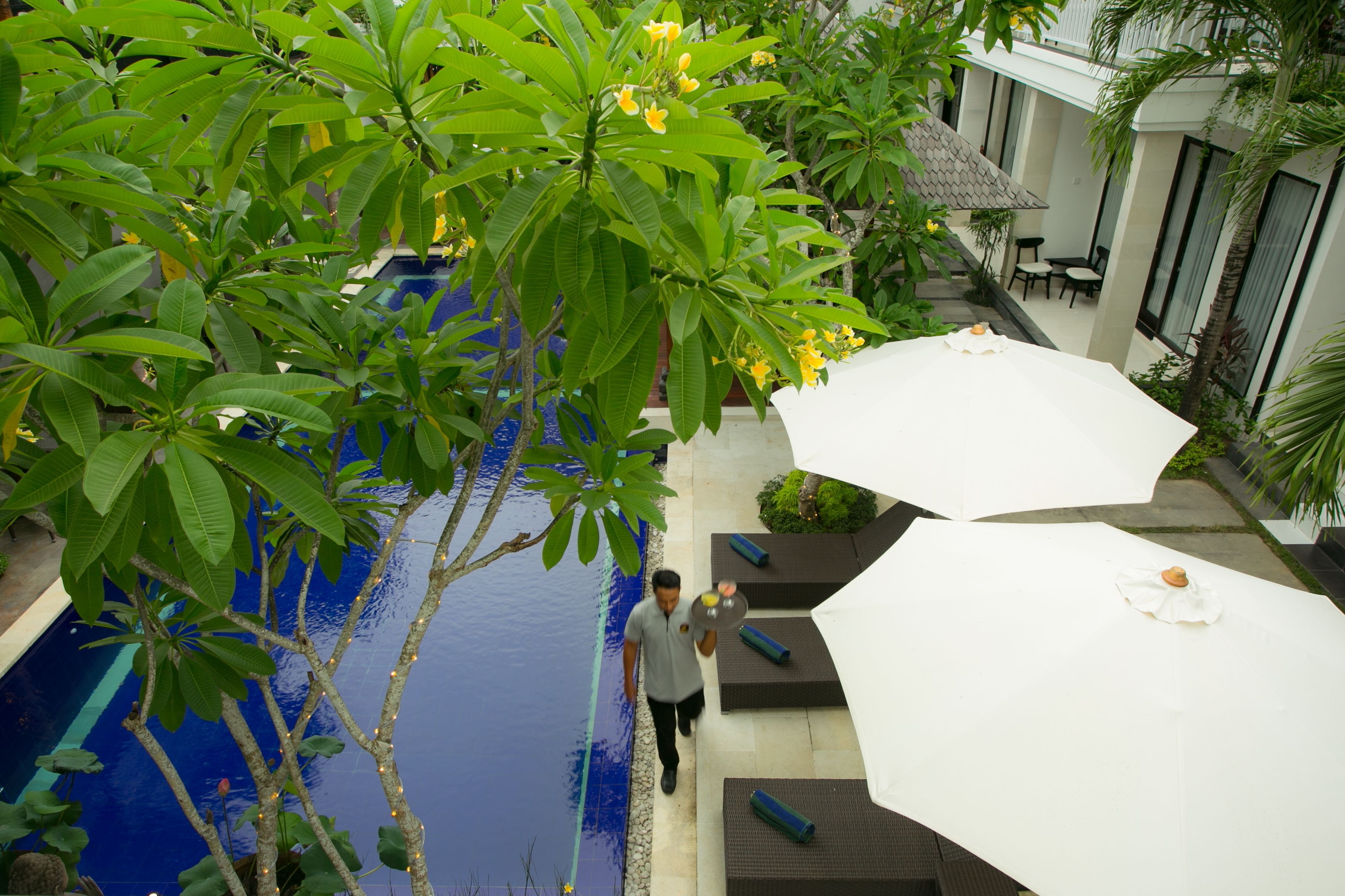 A poolside area with a man walking, surrounded by lush greenery and a white umbrella.
