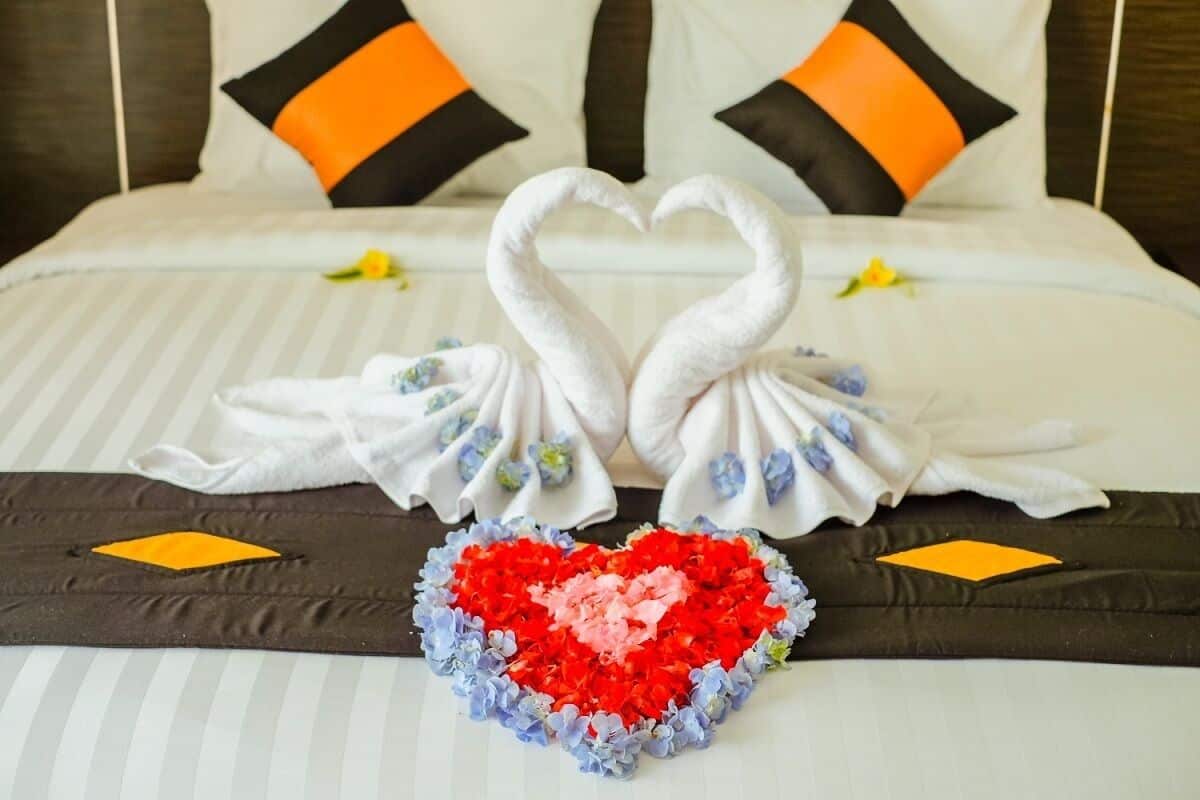 A bed with a heart-shaped arrangement of towels and flowers, two pillows with orange and black accents, and a striped bedspread.