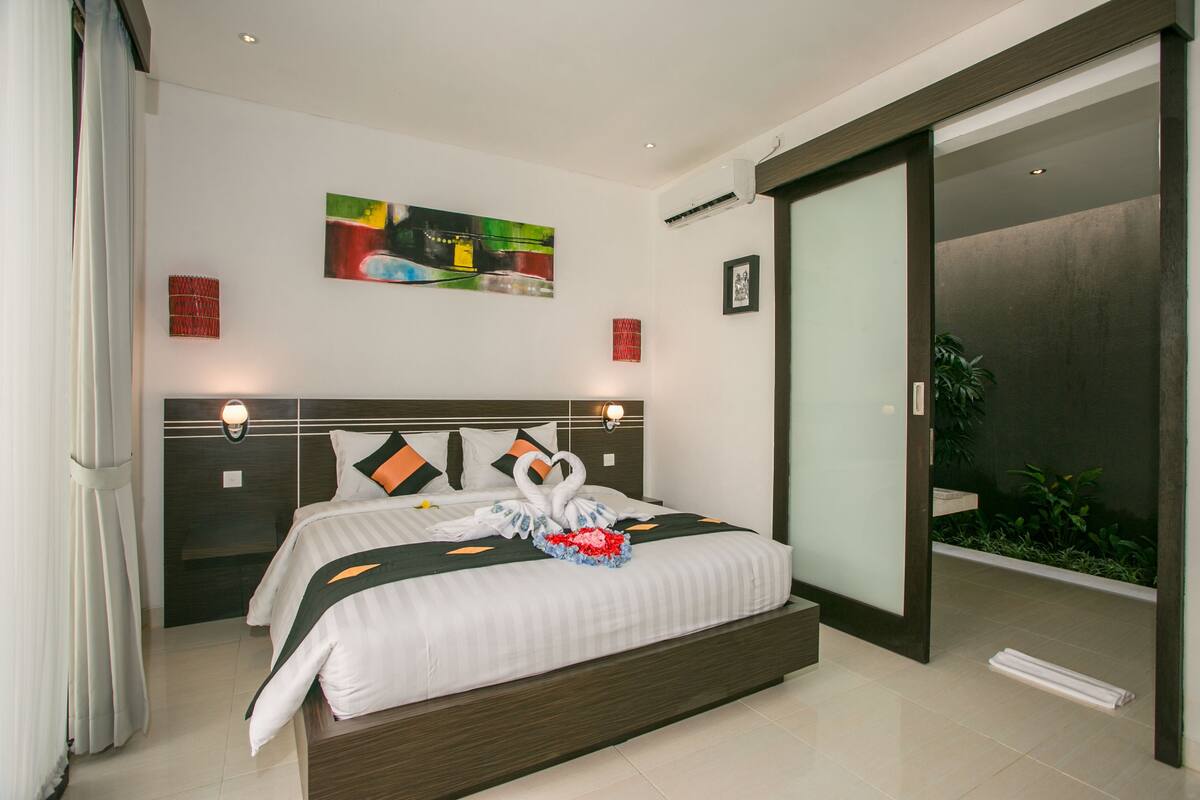 A modern hotel room with a large bed, a sliding glass door to a balcony, and a painting on the wall.