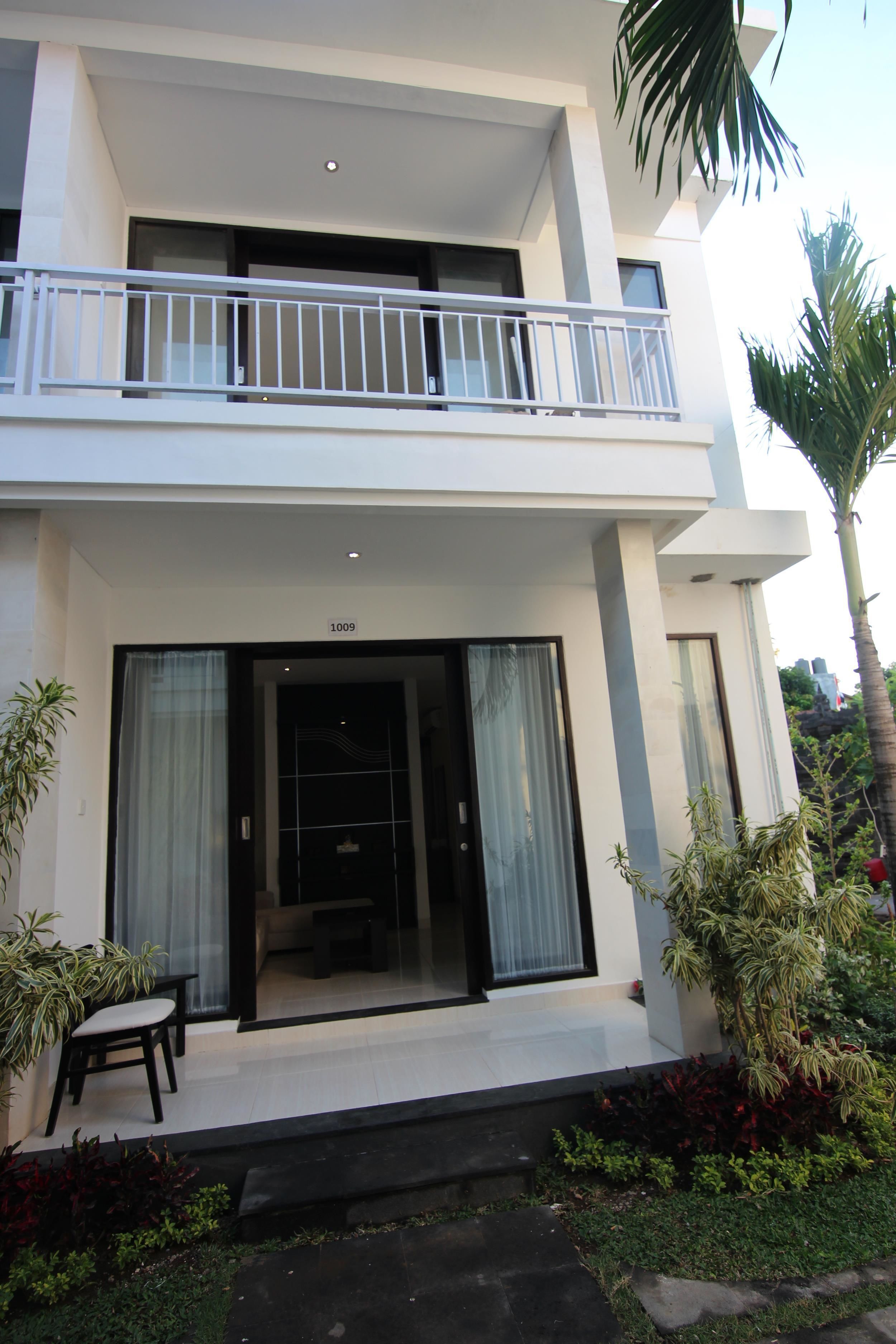 A modern two-story building with a balcony, a door with glass panels, and a small outdoor seating area.