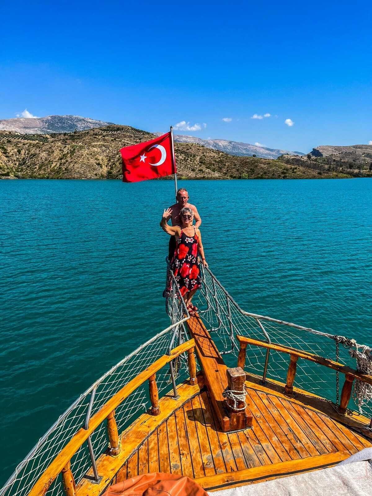 A boat deck with a Turkish flag, a person in a red floral dress, and a mountainous landscape in the background.