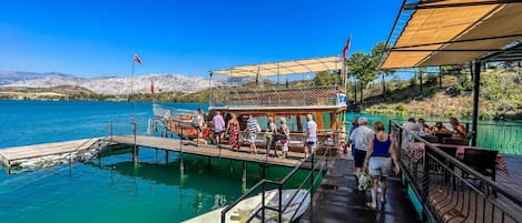 A boat docked at a pier with people boarding and disembarking, a covered seating area, and a view of the water and distant hills.