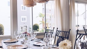 A dining area with a long table set for a meal, large windows, and a view of buildings and greenery outside. - Conny's Boutique Hotel - Adults Only (Manavgat)