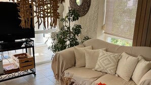 A living room with a beige sofa, a glass coffee table with decorative items, a television on a stand, and a large window with blinds. - Conny's Boutique Hotel - Adults Only (Manavgat)