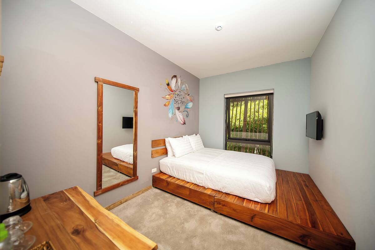A bedroom with a wooden bed, a large mirror, a TV, and a window with a view of greenery.