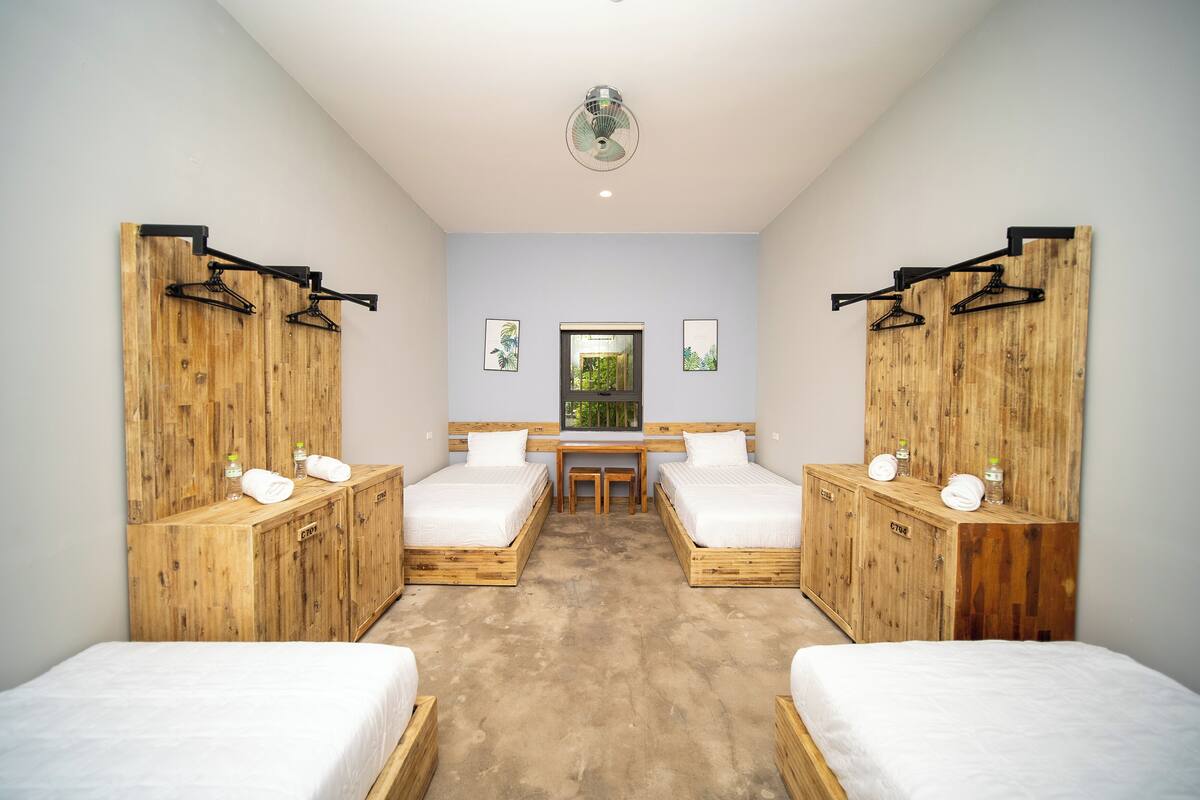 A room with two beds, a wooden wardrobe, a desk, and a window with a view of greenery.