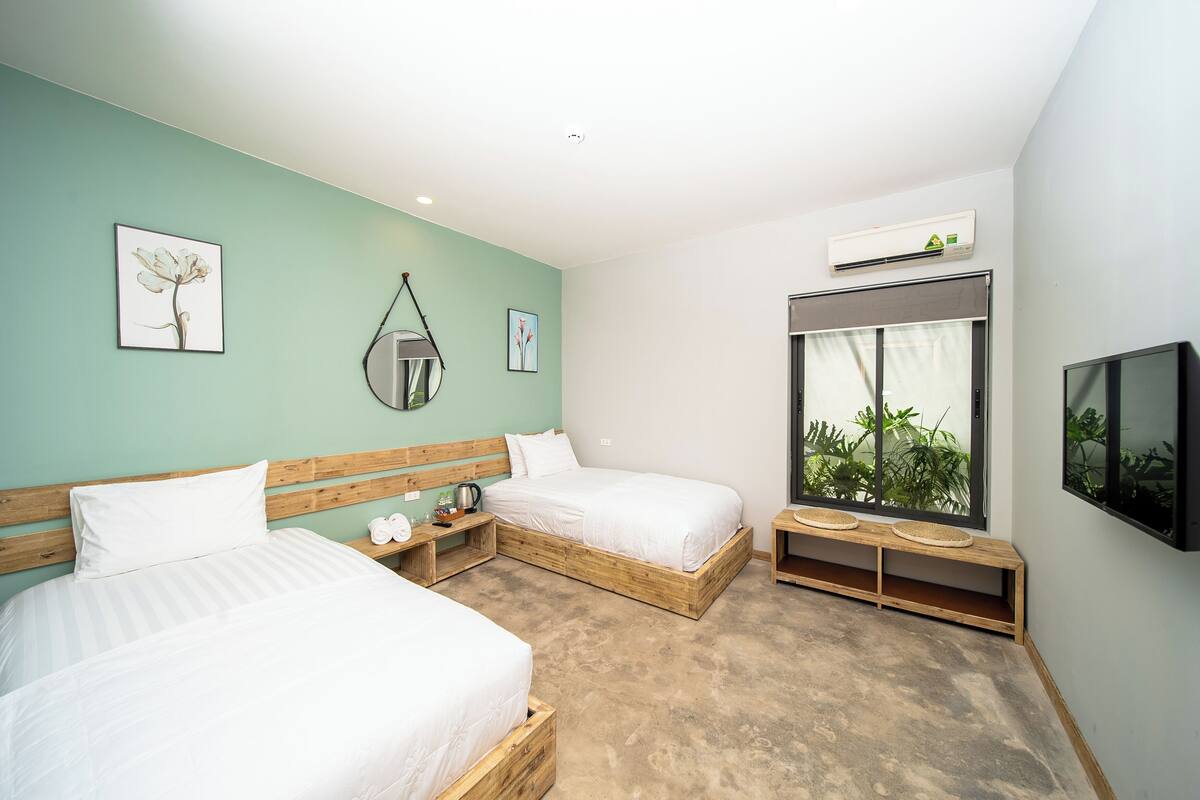 A modern bedroom with two beds, a wooden headboard, bedside tables, a bench, and a large window with a view of greenery.