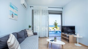 A modern living room with a sofa, a coffee table, a TV, and a view of a pool and beach. - Rhodes Sea Villas (Rhodes)