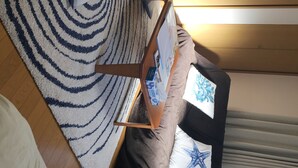 Una habitación con alfombra a rayas, un sofá con cojines, una mesita con una revista y una ventana con cortinas.