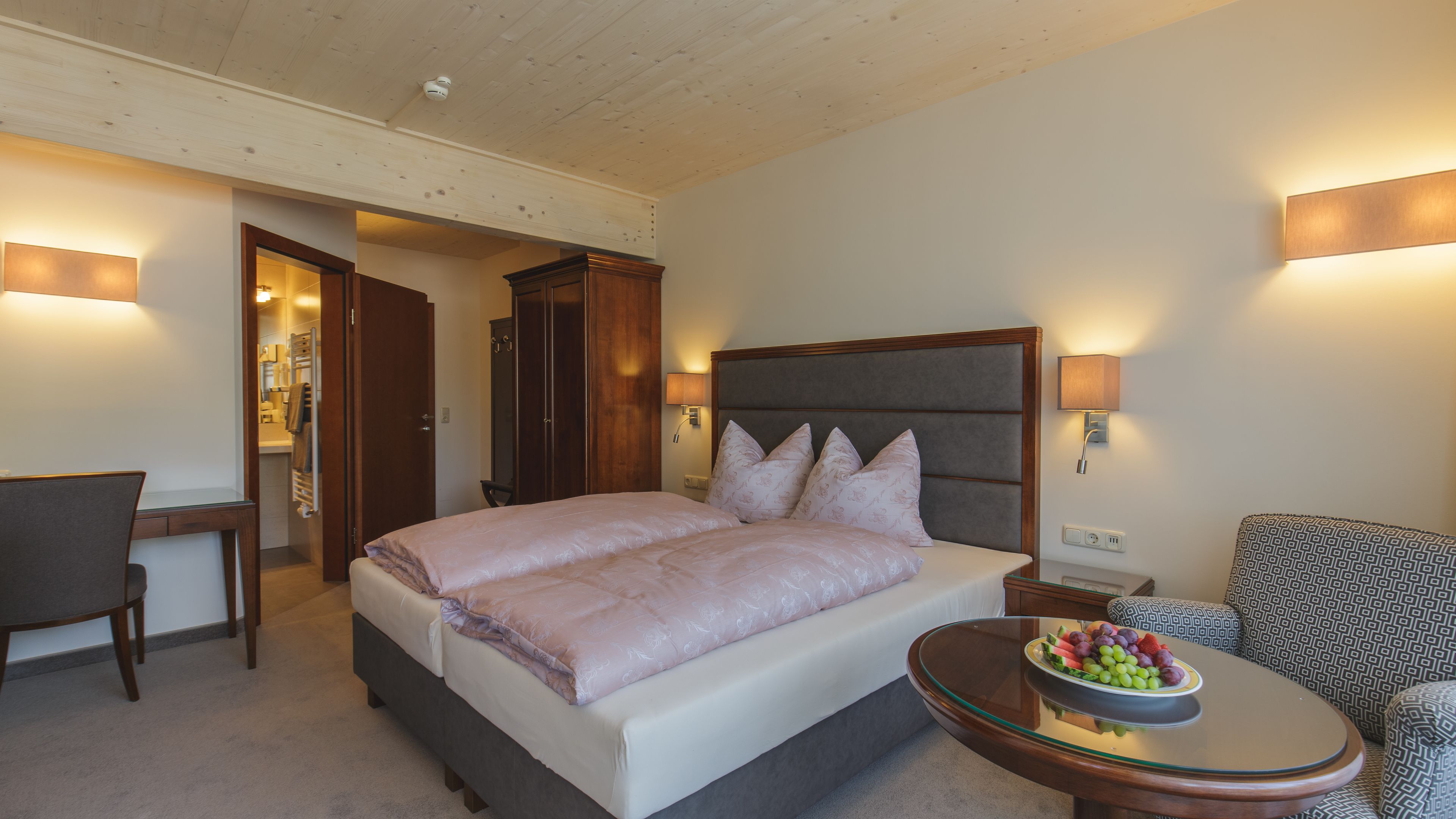 Ein Hotelzimmer mit Bett, Stuhl, einem kleinen Tisch mit Obst und einem durchgegangenen Türspalte sichtbaren Badezimmer.
