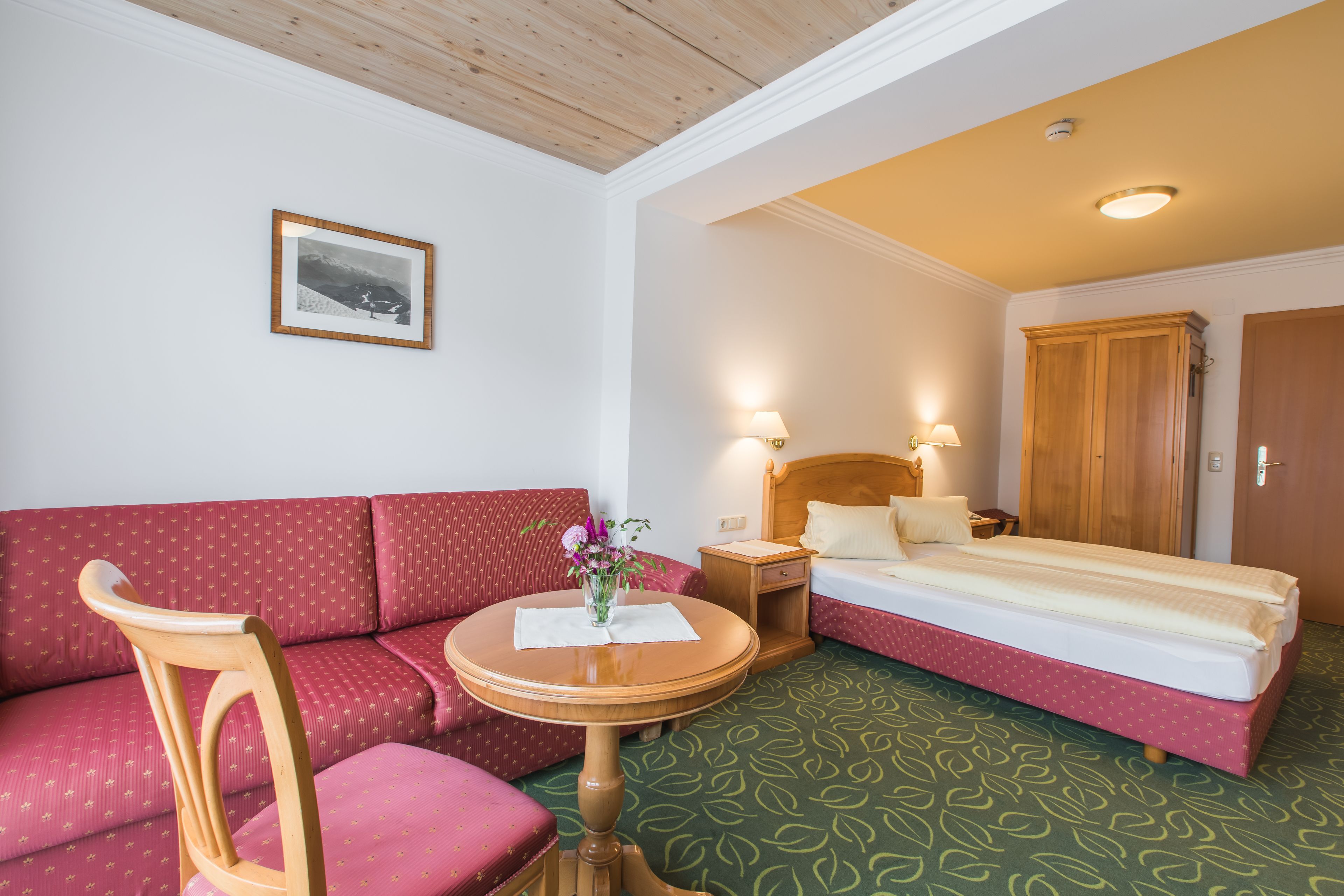 Een hotelkamer met een bed, een rode bank, een houten stoel, een kleine tafel met een vaas en een lijst met een foto aan de muur.