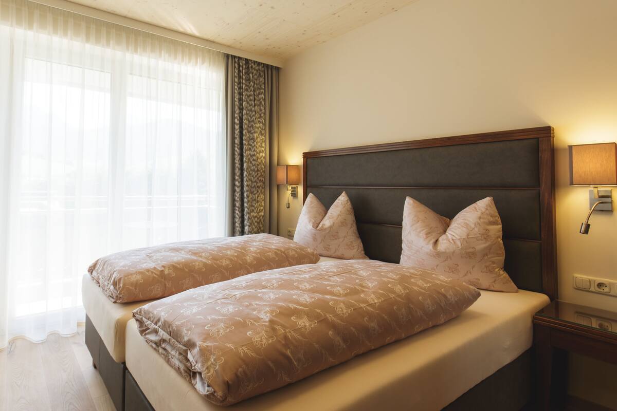 A hotel room with a double bed, a bedside table, a lamp, and a window with sheer curtains.