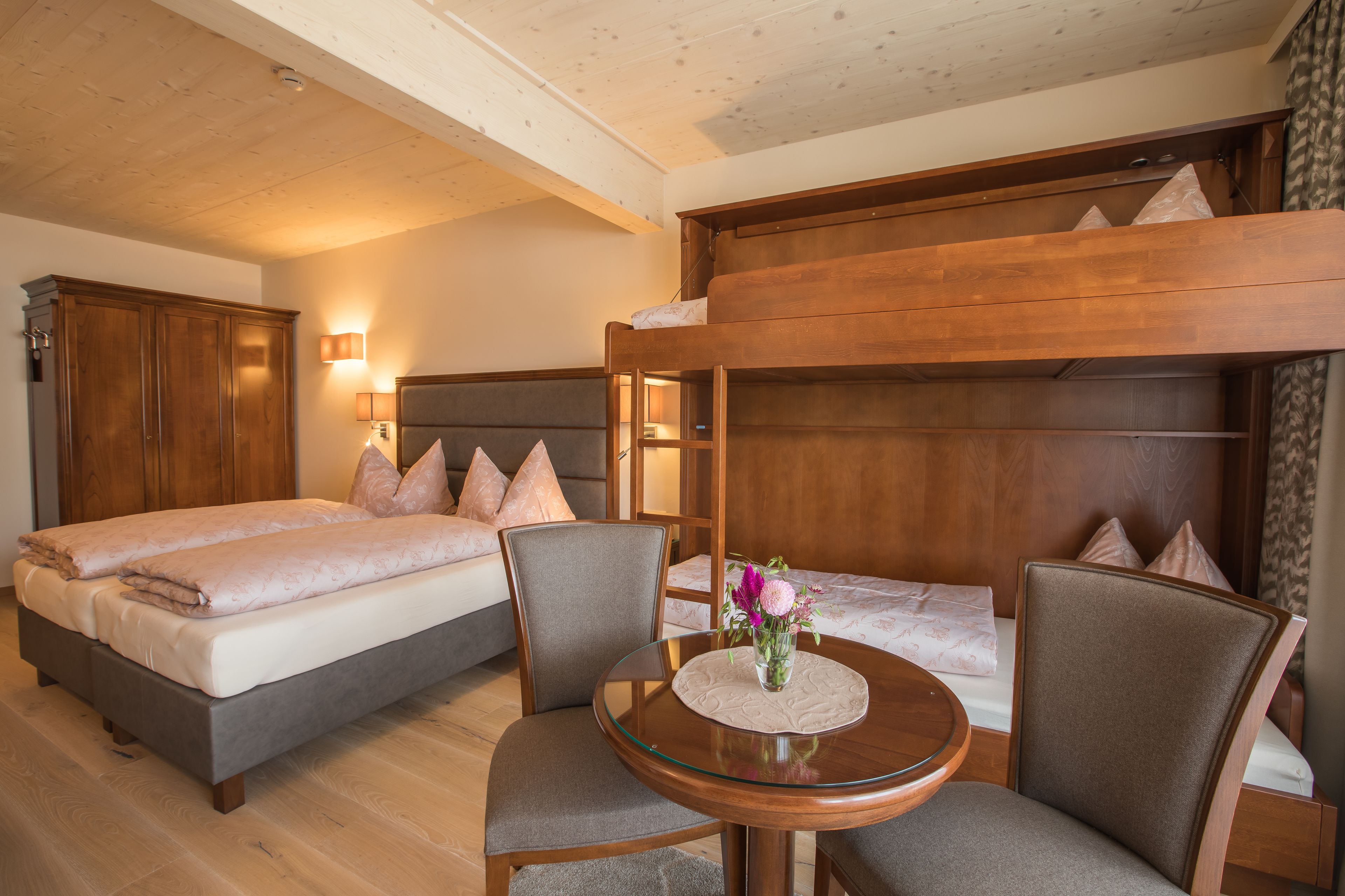 A hotel room with a bunk bed, a round glass table with a vase of flowers, and a wooden wardrobe.