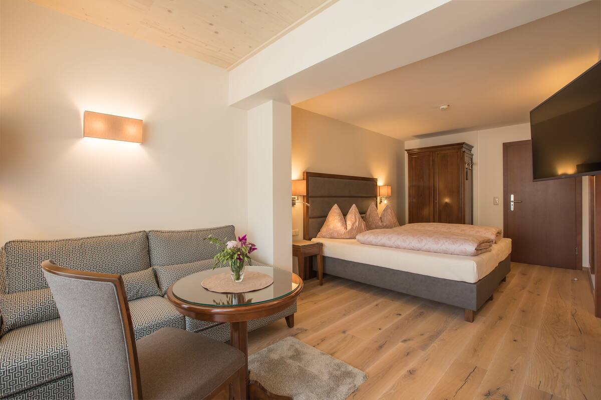 A modern bedroom with a bed, a sofa, a small table with a vase, and a TV mounted on the wall.