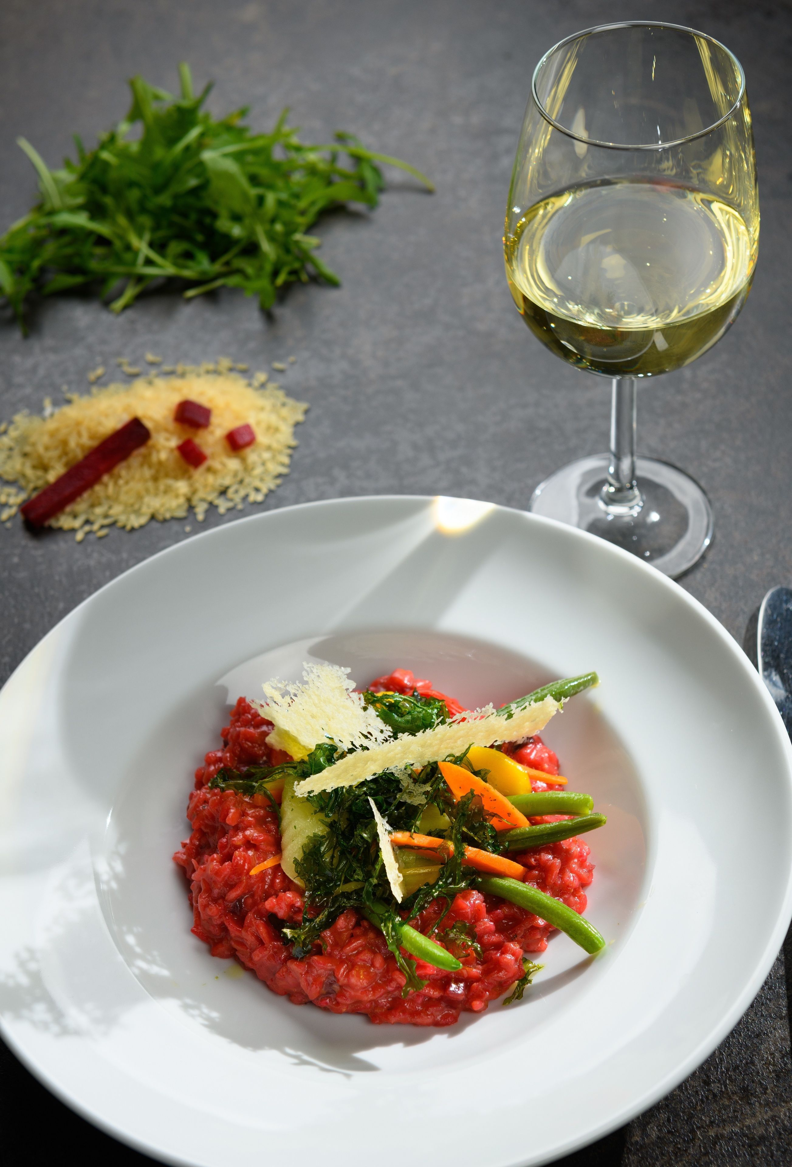 A white plate with a dish of red risotto, garnished with green vegetables and cheese, accompanied by a glass of white wine on a dark surface.