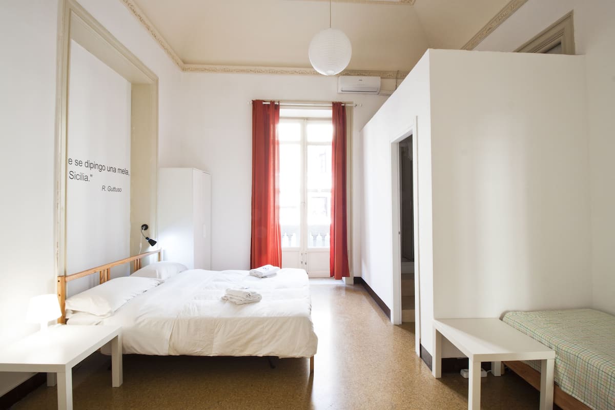 A hotel room with two beds, a desk, a wardrobe, and a window with red curtains.