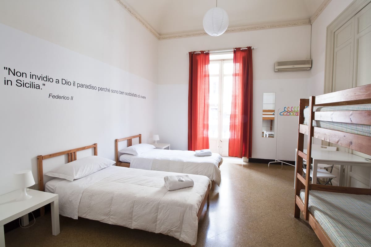 A hotel room with two single beds, a desk, a chair, a wardrobe, and a window with red curtains.