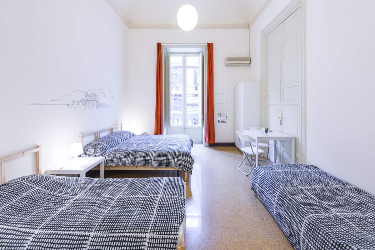 A hotel room with two single beds, a desk, a chair, and a large window with curtains.
