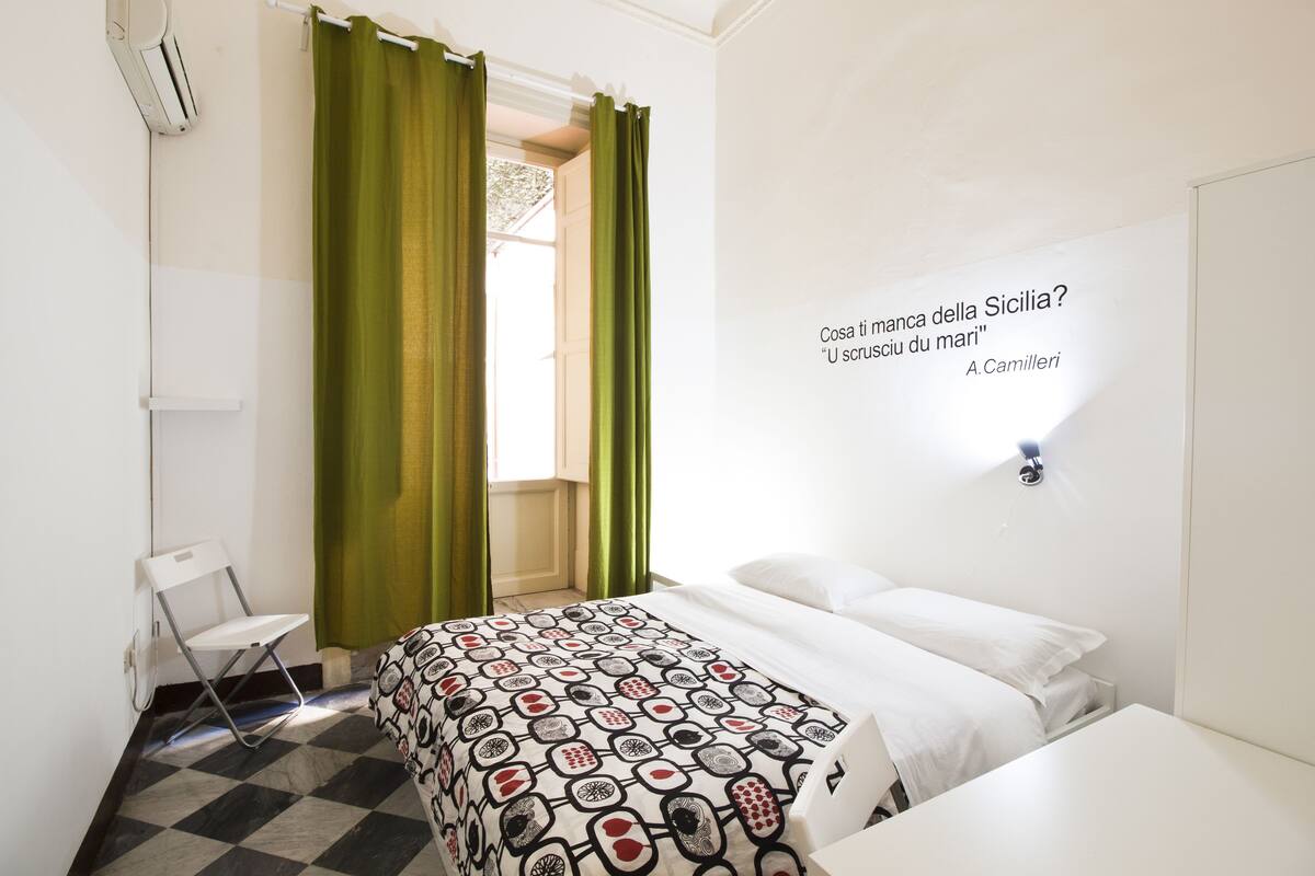 A bedroom with a bed, a chair, a wall-mounted light, and a window with green curtains.