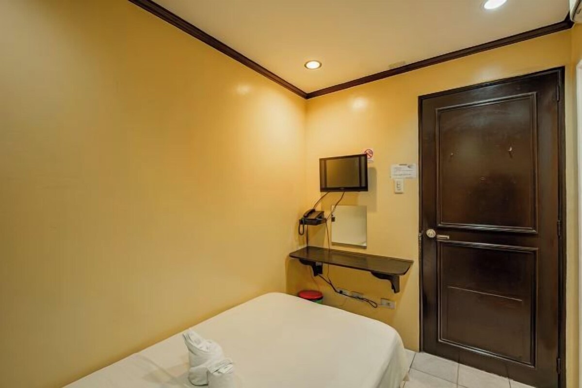 A small, single-bed room with a bedside table, a television mounted on the wall, and a door with a handle.