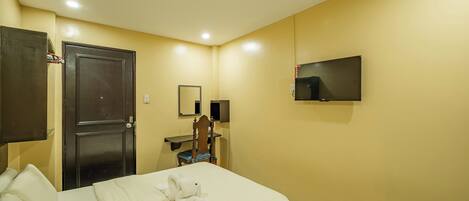 Quarto de hotel com uma cama, escrivaninha, cadeira, televisão e guarda-roupa.