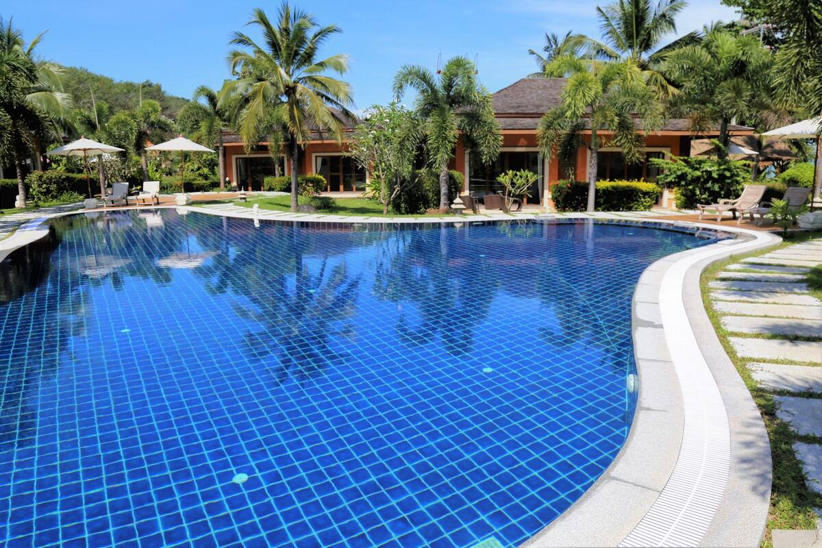 Une grande piscine d’un bleu éclatant, bordée de carreaux, entourée de palmiers et de chaises longues.